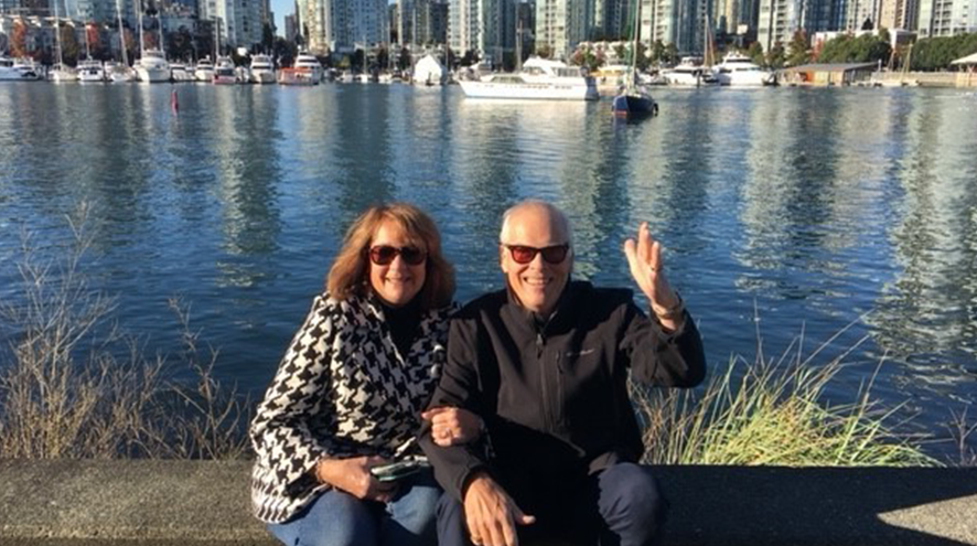 A woman with long sandy blonde hair sits next to a man with white hair, waving. They are seated on a bench in front of water and with a city on the other side.