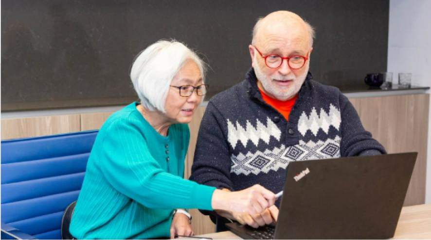 two people wearing glasses, smiling and looking at their computer screen