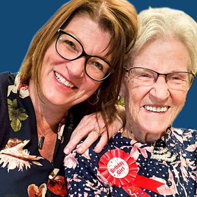 A middle-aged woman with long brown hair cuddles up with an older woman with a badge reading "Birthday girl" on her lapel.