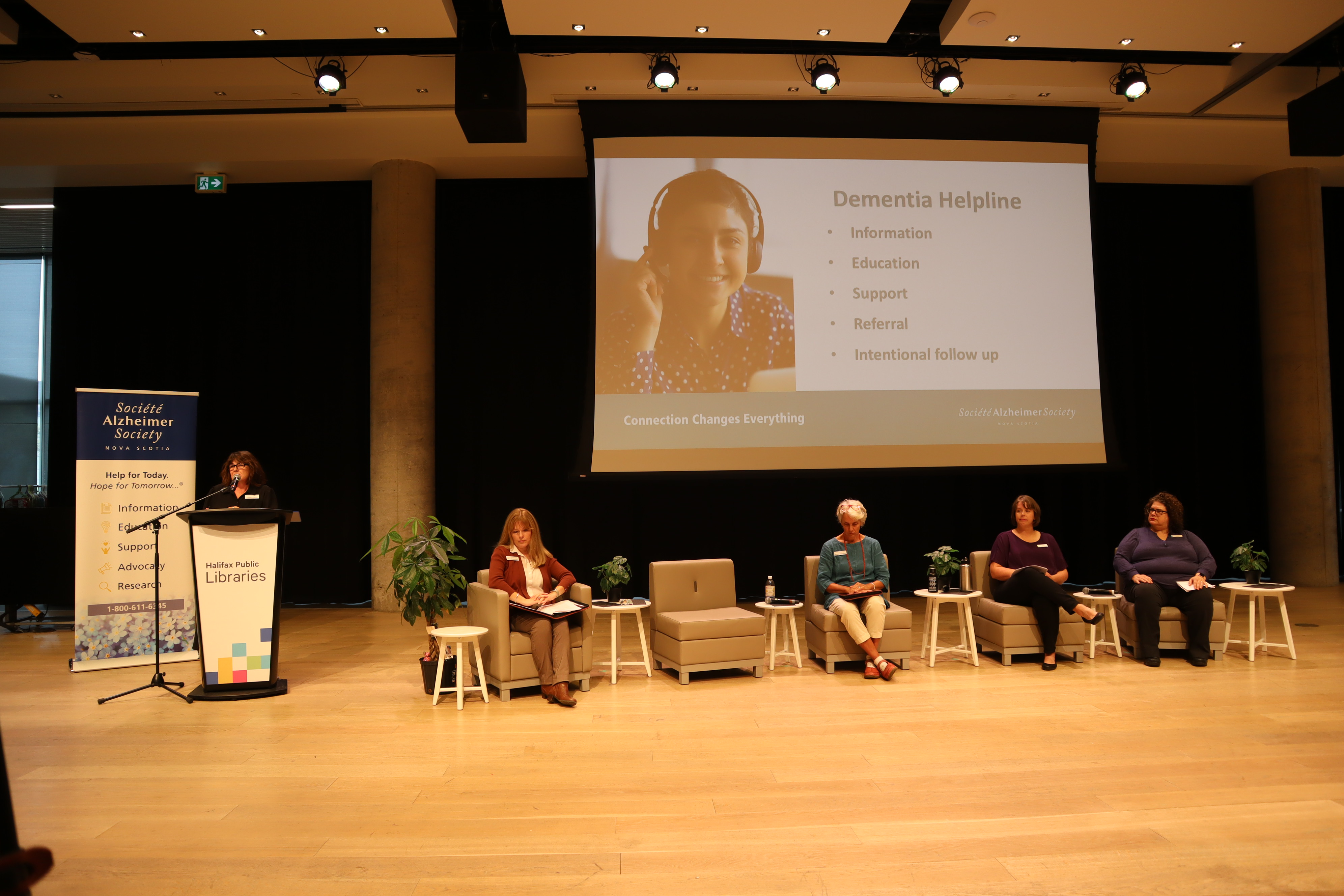 A speaker stands at a podium to the left. 4 other speakers are seated in chairs to their right. A big projector screen is displayed behind them with information about ASNS programs and services.