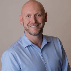 John wears a blue button down shirt smiling at the camera.