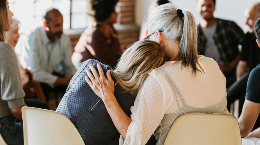 Two women embracing shown from behind as they participate in a group counselling session, which is blurred in the background..