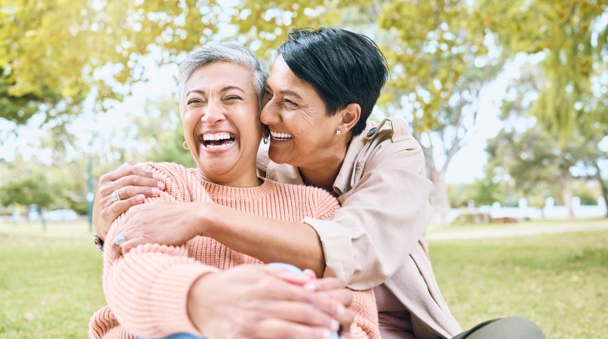 Older couple, both with light olive skin, laughing and embracing outdoors. One of the women is wearing a pink sweater and has short silver hair, and the other has short dark hair and is wearing a light-coloured jacket.