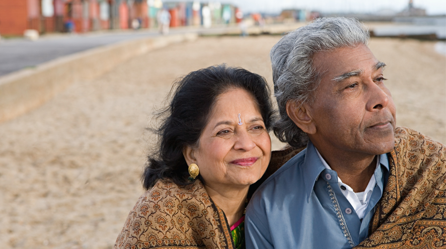 Older woman and older man sit closely wrapped in a shawl at the beach