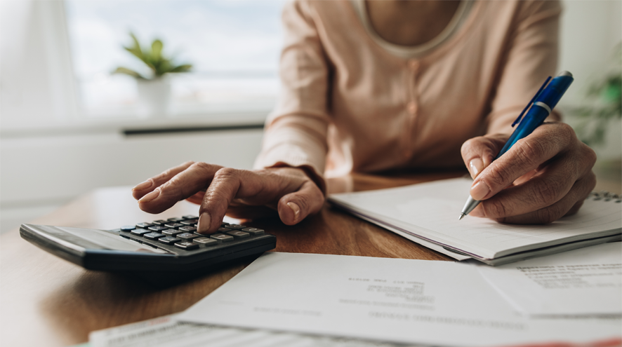 A woman sits at a table with a calculator, paper, pen and bills