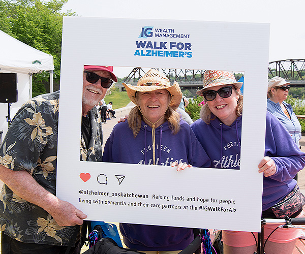 A husband, wife, and adult daughter standing behind an over-sized white photo frame that says "IG Wealth Management Walk for Alzheimer's"