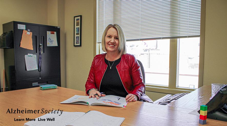 Joanne Bracken sitting at her desk with a day planner. She has blonde hair, is wearing a bright red jacket, and is smiling.