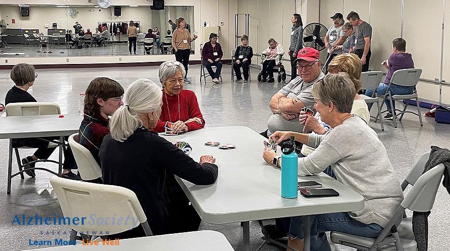 A group of people smiling and playing a card game, while another group of people are doing physical activity.