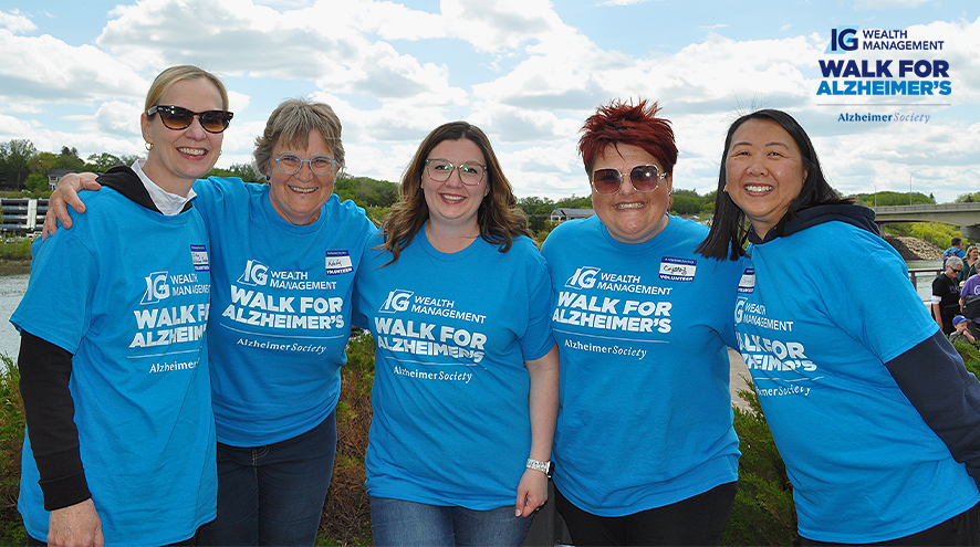 Five women wearing light blue IG Wealth Management Walk for Alzheimer's t-shirts. They are standing in front of Saskatoon's River Landing. The sun is shining and the leaves are bright green.