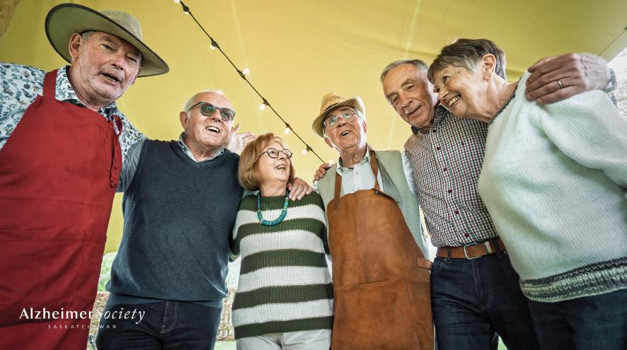 A group of people smiling with their arms around each other at on outdoor cookout. Some are wearing aprons.