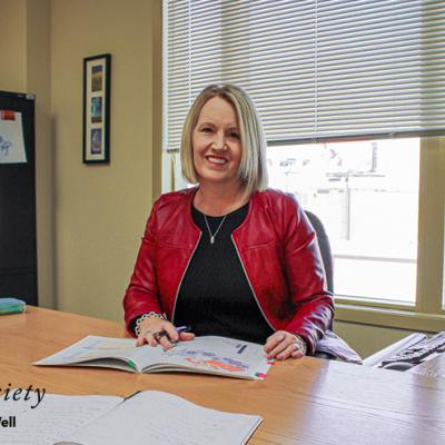 Joanne Bracken sitting at her desk with a day planner. She has blonde hair, is wearing a bright red jacket, and is smiling.