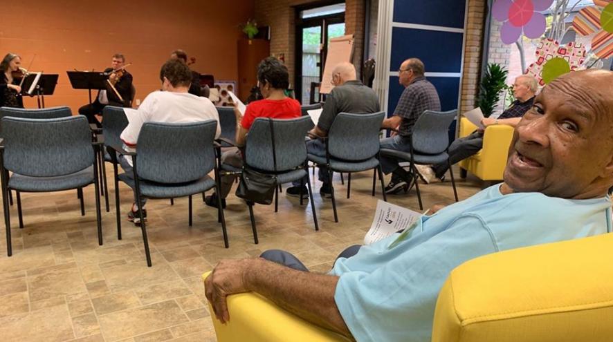 Photo of a group of people sitting in a room listening to a small orchestra.