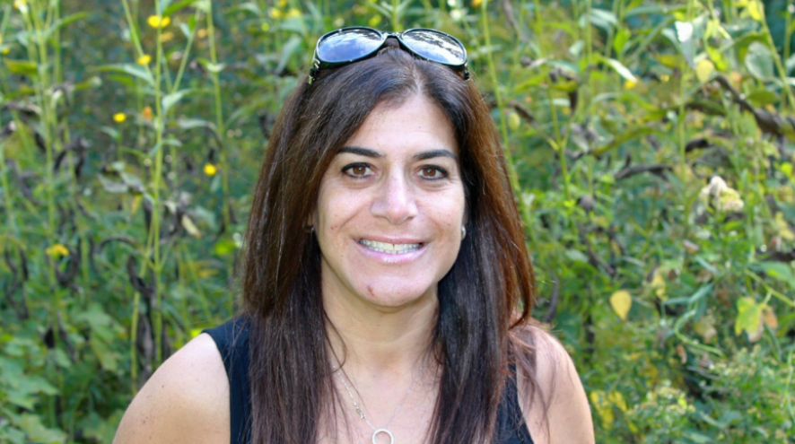 Head and shoulder shot of Bonnie Wisener, a registered holistic nutritionist, sitting in front of tall green plants some with yellow flowers.