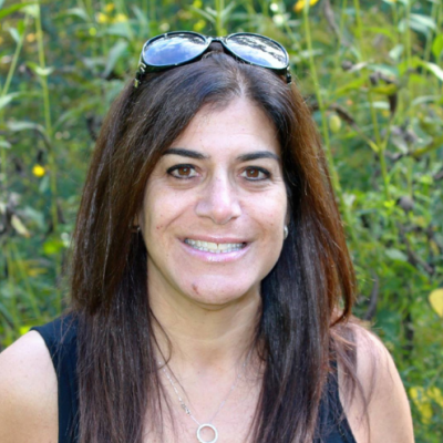 Head and shoulder shot of Bonnie Wisener, a registered holistic nutritionist, sitting in front of tall green plants some with yellow flowers.