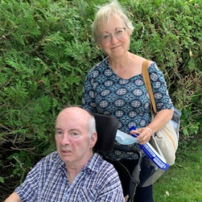 Older woman with short hair standing behind an older man in a wheelchair outside.